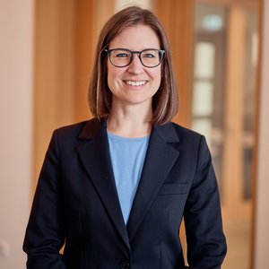 Frau mit braunen Haaren und Brille in dunkelblauem Sacko in einem hellen Raum schaut offen lächelnd in die Kamera