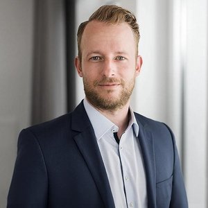 Dunkelblonder Mann in blauem Sacko mit hellblauem Hemd blickt der Kamera leicht zugedreht offen lächend in die Kamera.