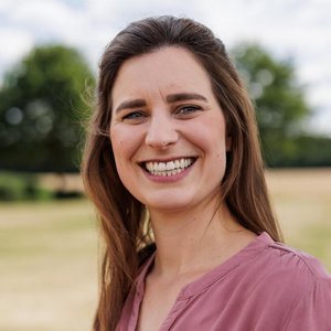 Eine jungte Frau mit offenen, lange, braunen Haaren, schaut offen lachend leicht von der Seite in die Kamera. Im Hintergrund sind Bäume zu sehen. Sie trägt ein blassrotes Hemd aus Baumwolle oder Leinen.